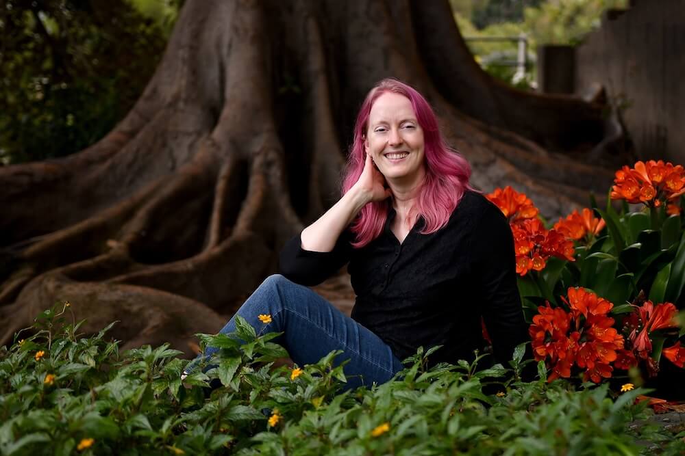 Woman with pink hair sits amidst greenery, reflecting on childhood trauma and attachment wounds.