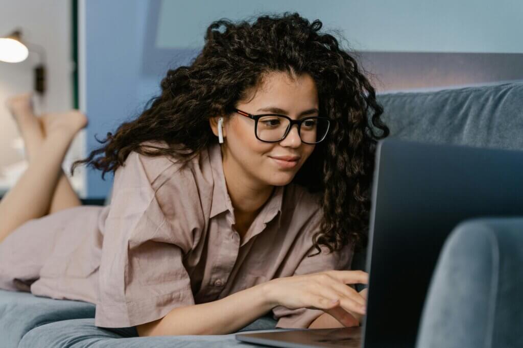 Woman relaxing on couch, using laptop, wearing glasses and earbuds. Social media impact on mental health.